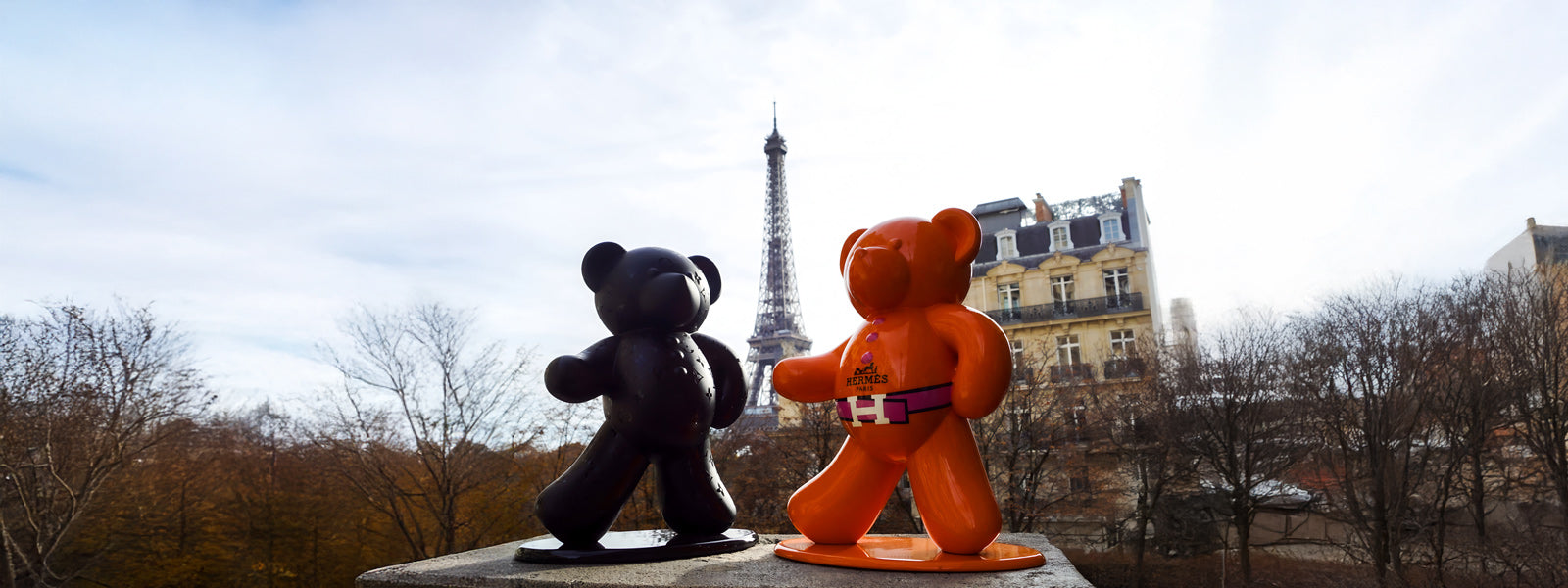 A person with a blue bear head balloon standing near a Parisian street with the Eiffel Tower in the background. They hold a green bear balloon and wear a white shirt.