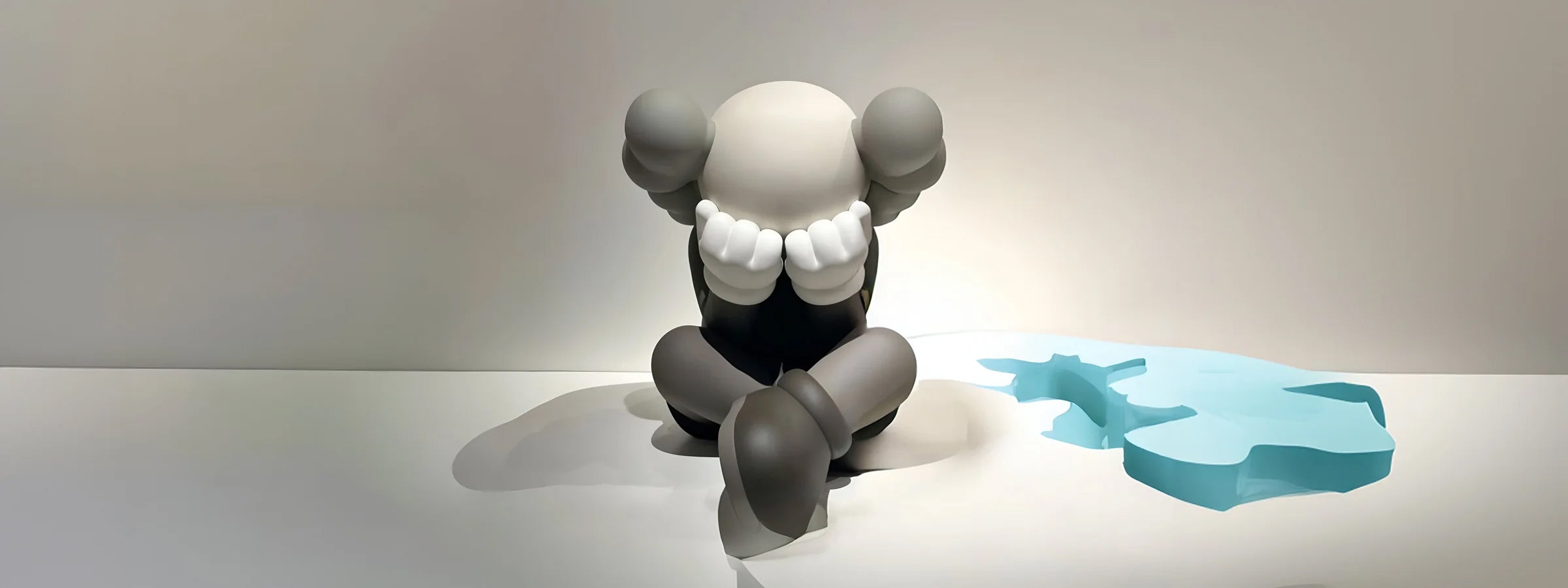 Gray and white abstract iconify sculpture with large round ears, sitting with its head in its hands. The sculpture has exaggerated limbs and is placed on a white surface beside a small blue puddle, evoking a sense of sadness or contemplation.