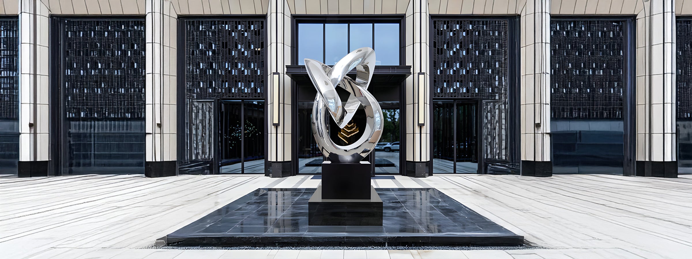 Large polished stainless steel abstract sculpture with interlocking loops, displayed on a black pedestal in an outdoor plaza with modern architecture and reflective black flooring.