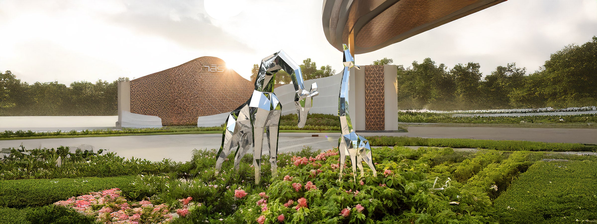 Two stainless steel geometric giraffe sculptures, one large and one small, standing in a lush garden with pink flowers. The sculptures have a reflective, faceted design, set against a modern architectural backdrop with a large curved structure.