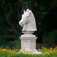 The 120cm Noble Horse Head White Marble Outdoor Sculpture by Giant Sculptures stands on a pedestal amidst lush greenery, with yellow flowers at its base enhancing the vibrant contrast of this exquisite equestrian artwork.