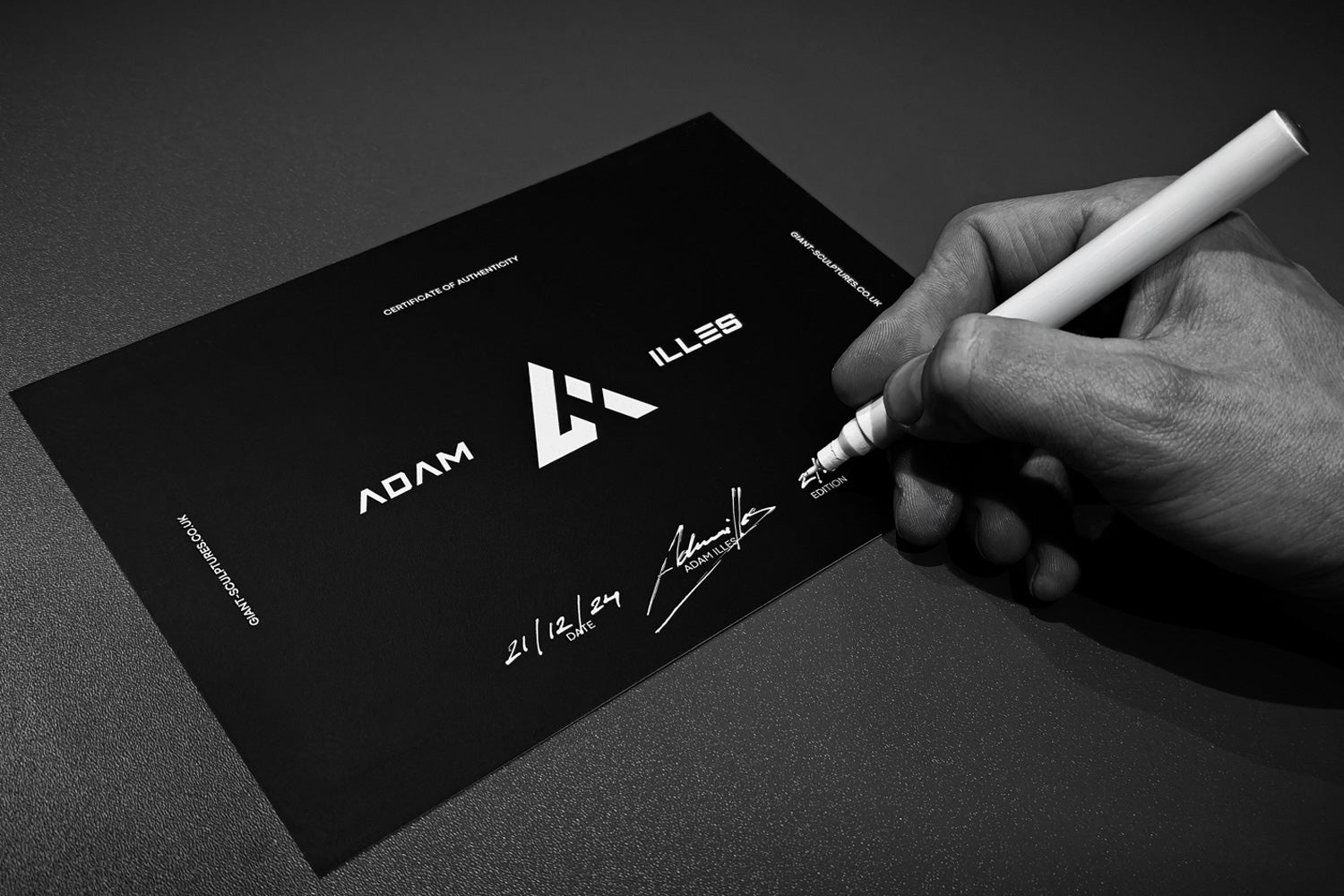 A hand holds a white marker pen, signing a certificate labeled Certificate of Ownership for Adam Illes. The logo and signature are prominent on a dark surface. The date 21.10.19 is handwritten near the signature.