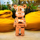 The Rose Gold Bear Sculpture - 50cm by Giant Sculptures, with its shiny, metallic finish, is positioned on a floor amidst vibrant yellow bean bags and lush green potted plants. This arrangement creates a modern and eclectic indoor setting that celebrates contemporary artistry.