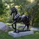 The Giant Sculptures Graceful Horse Black Marble Outdoor Sculpture - 220cm stands elegantly on a stone pedestal amidst the verdant garden. Sunlight filters through the trees, casting dappled shadows on the grass and nearby rocks, giving the dynamic horse sculpture an appearance of being in mid-stride.