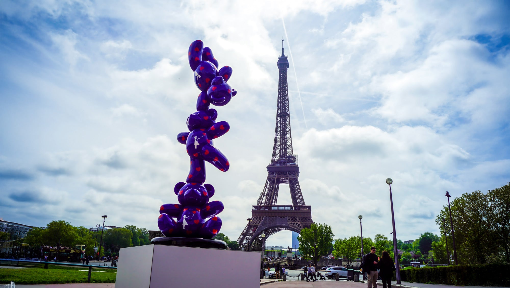 A colorful balloon animal sculpture on a pedestal stands prominently in front of the Eiffel Tower under a partly cloudy sky. The sculpture is purple with red and orange accents, contrasting with the iconic Paris landmark in the background.