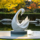 The Graceful Swan White Marble Outdoor Sculpture by Giant Sculptures, standing at 100cm, sits elegantly on a pedestal surrounded by water. Lush greenery and vibrant autumn foliage enhance the serene and picturesque outdoor setting.