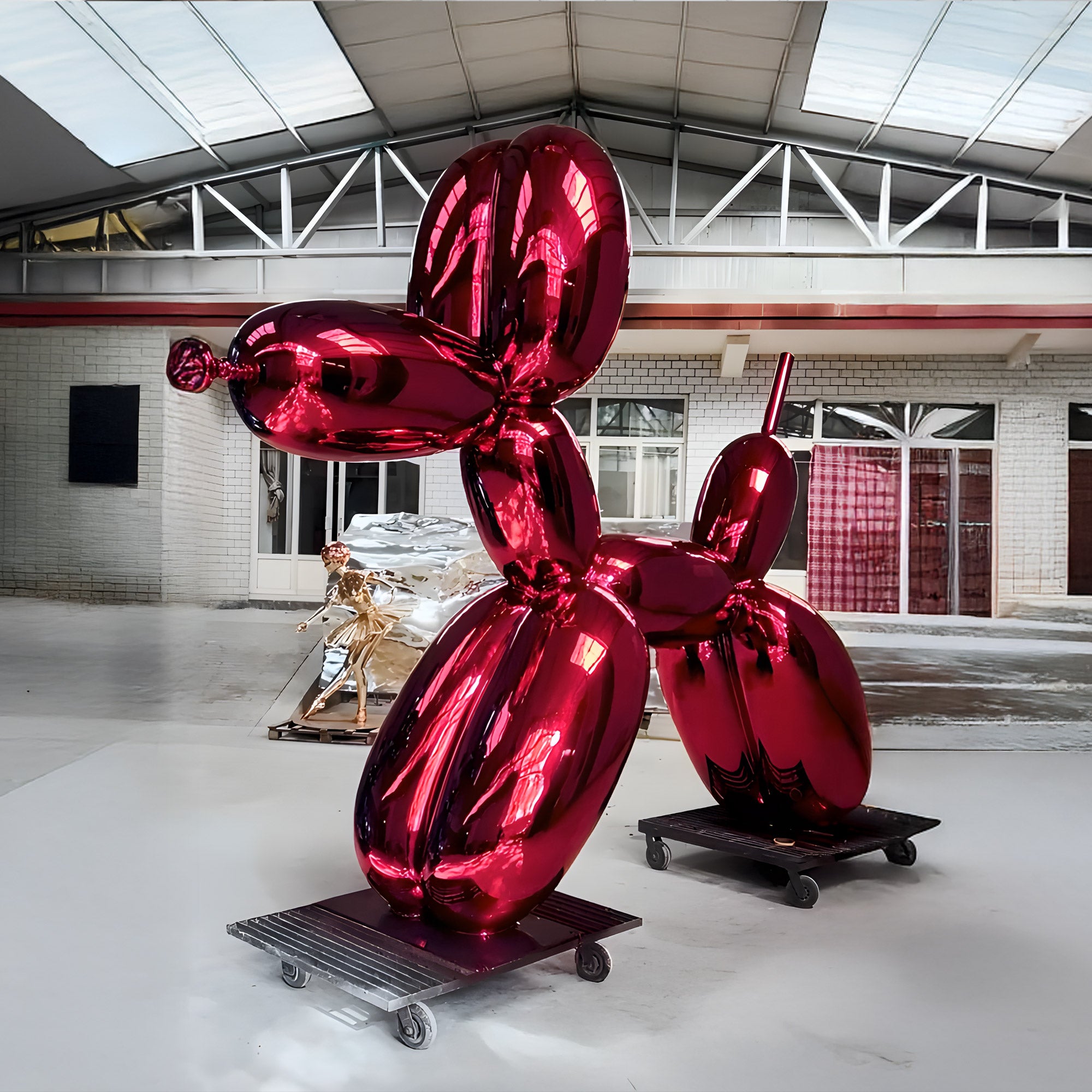 A large, glossy red balloon dog sculpture stands indoors on a wheeled platform. The background features a warehouse-style space with metal beams and a smaller golden sculpture in a similar style.