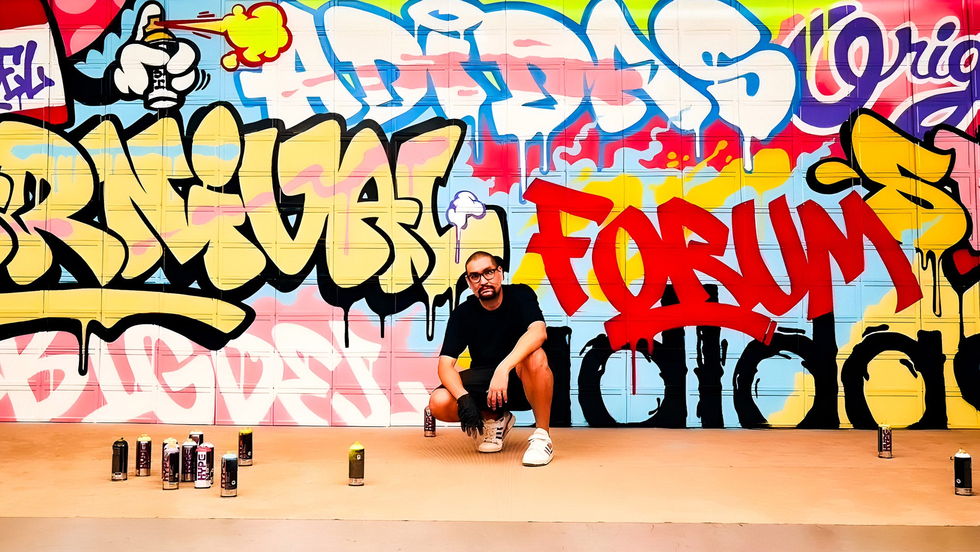 A person crouches in front of a colorful graffiti wall featuring words like FORUM and various tags. Several spray paint cans are scattered on the ground. The person is wearing a black shirt and shorts, and the scene is vibrant and artistic.