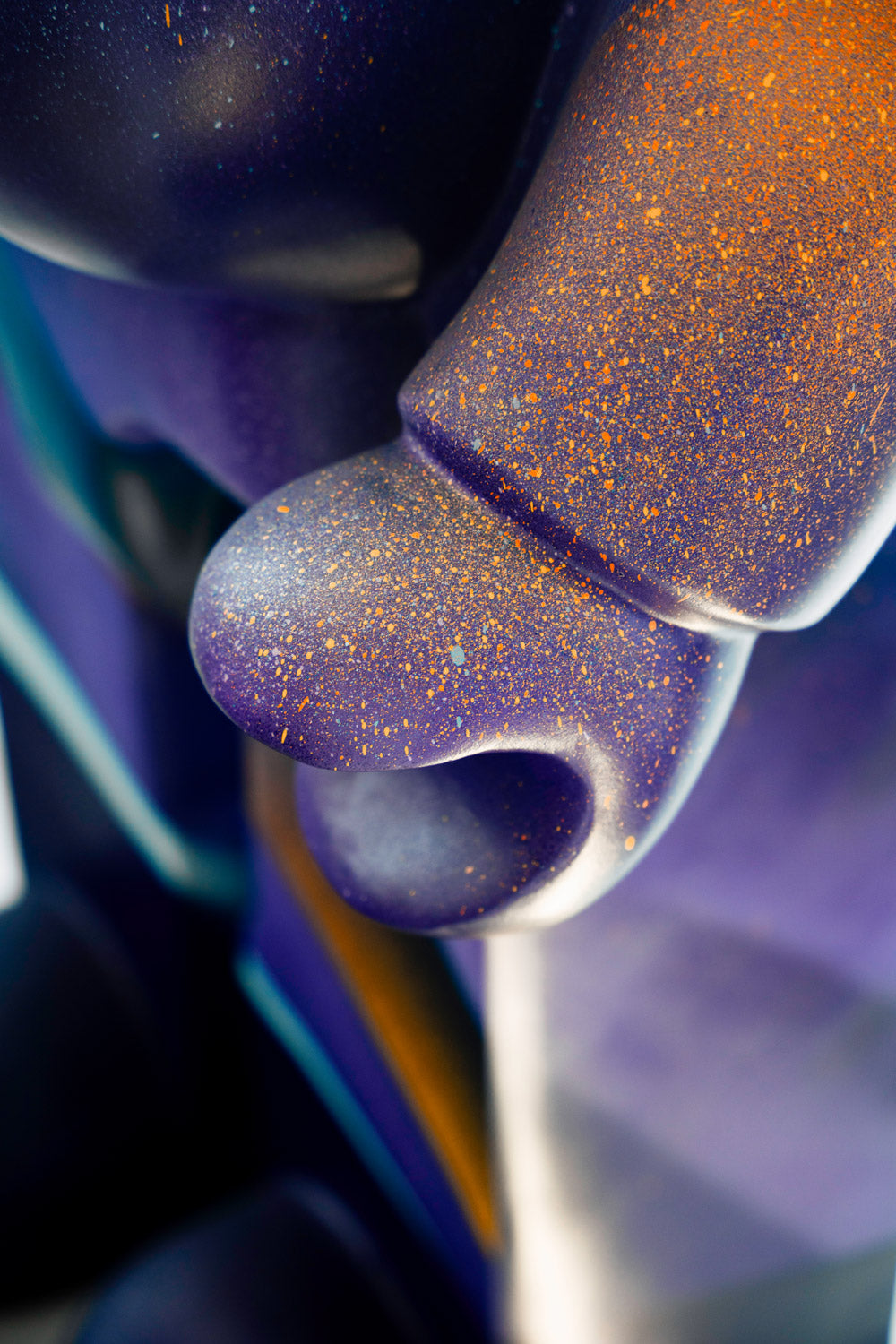 Close-up of a colorful, abstract sculpture with smooth, curved surfaces. The surface features a gradient of blue and purple with a speckled pattern of orange and white, creating a shimmering effect.