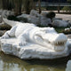 The Crocodile III 60cm white marble sculpture by Giant Sculptures rests on a rocky platform, encircled by a pond. Trees and shrubs enhance the serene outdoor setting of this detailed work.