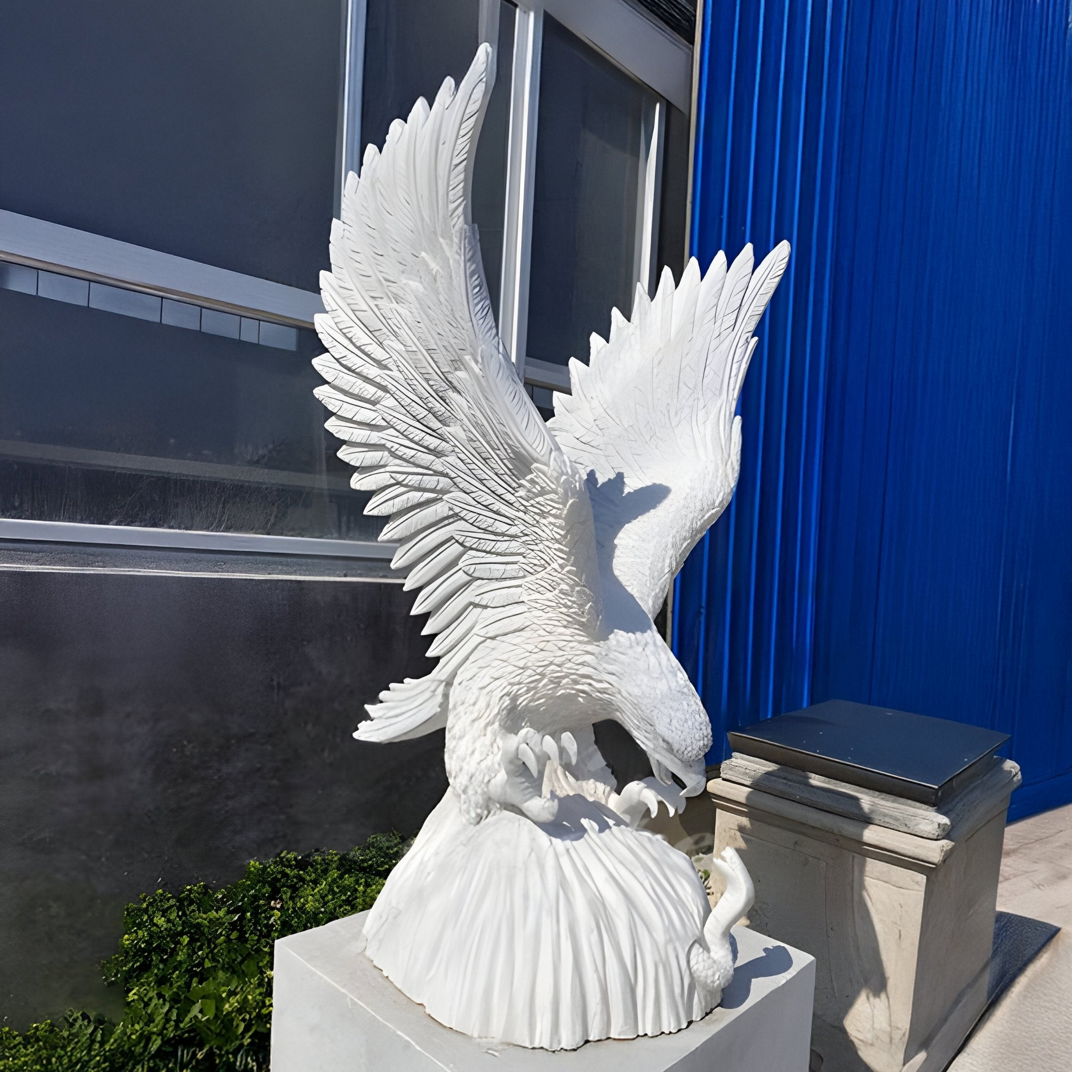 The Soaring Eagle White Marble Outdoor Sculpture by Giant Sculptures, standing 100cm tall, displays a magnificent eagle on a rectangular pedestal with spread wings and talons gripping prey. The scene includes a modern building with glass and blue panels, accented by greenery below.