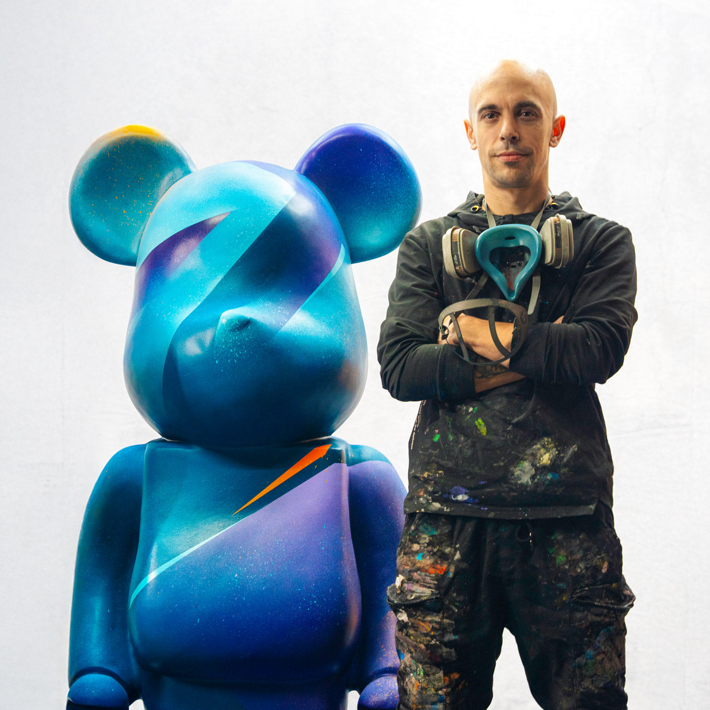 A person stands with arms crossed beside a large, colorful bear sculpture. The sculpture is primarily blue with abstract patterns. The person is wearing black clothing with paint stains and has a respirator around their neck.