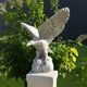 The Giant Sculptures Guardian Wings Eagle Marble Outdoor Sculpture, standing 120cm tall, perches majestically on a pedestal amidst lush green foliage. Sunlight casts enchanting shadows over the outdoor scene.