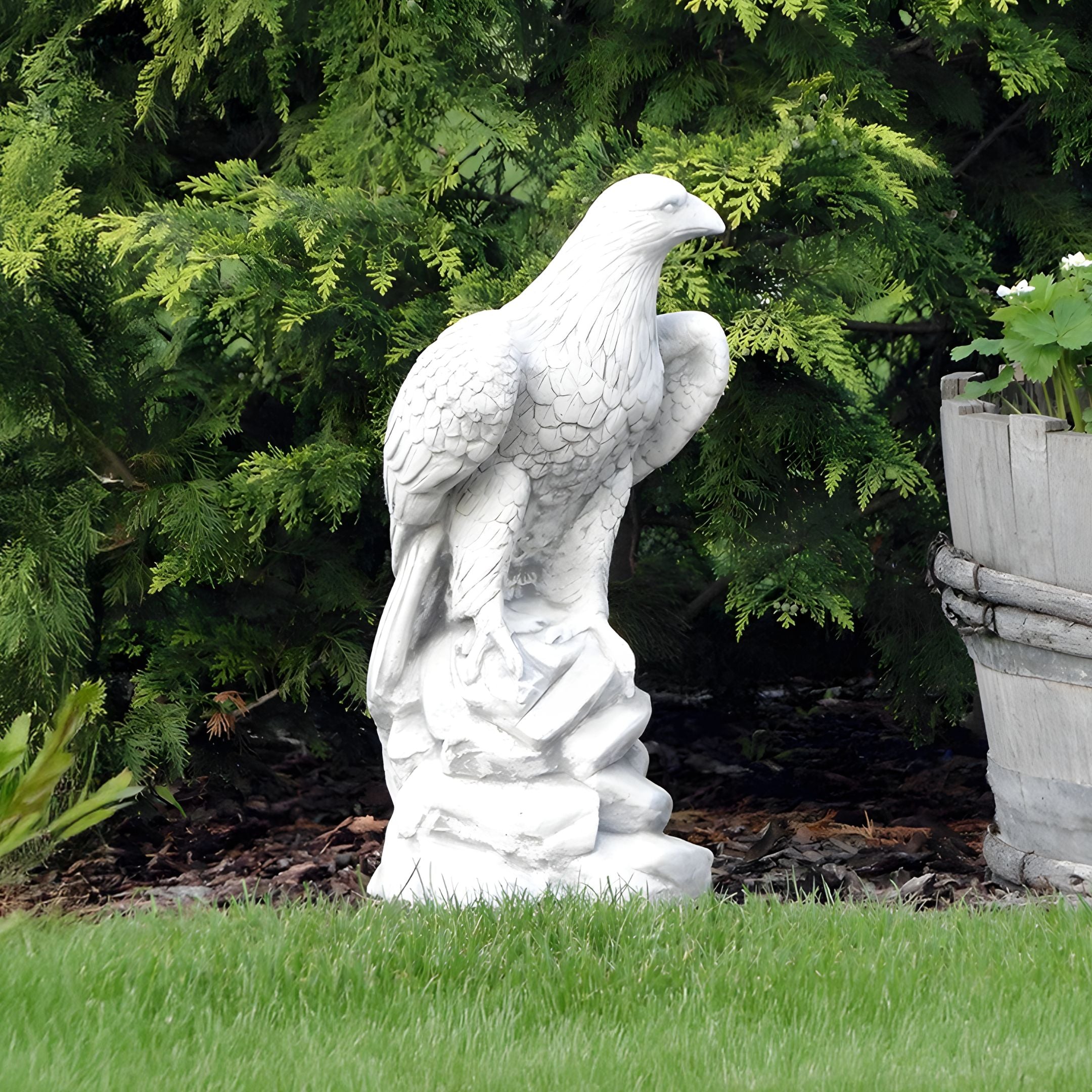 In a garden setting, the Regal Eagle White Marble Outdoor Sculpture - 120cm by Giant Sculptures stands on a rocky base, surrounded by lush plants and a wooden planter.