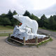 The Polar Bear Family White Marble Outdoor Sculptures (180cm) by Giant Sculptures features a large bear standing with a smaller one on a rocky base, surrounded by wooden posts in an outdoor park setting, capturing nature-inspired garden art amidst trees and a road for serenity.