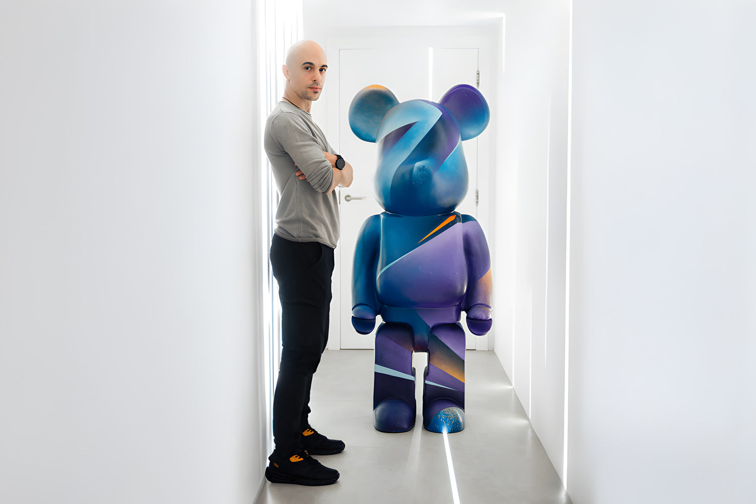 A person standing in a minimalist white room with arms crossed, posing next to a large, artistic bear-shaped figure painted in shades of blue and purple with abstract patterns.