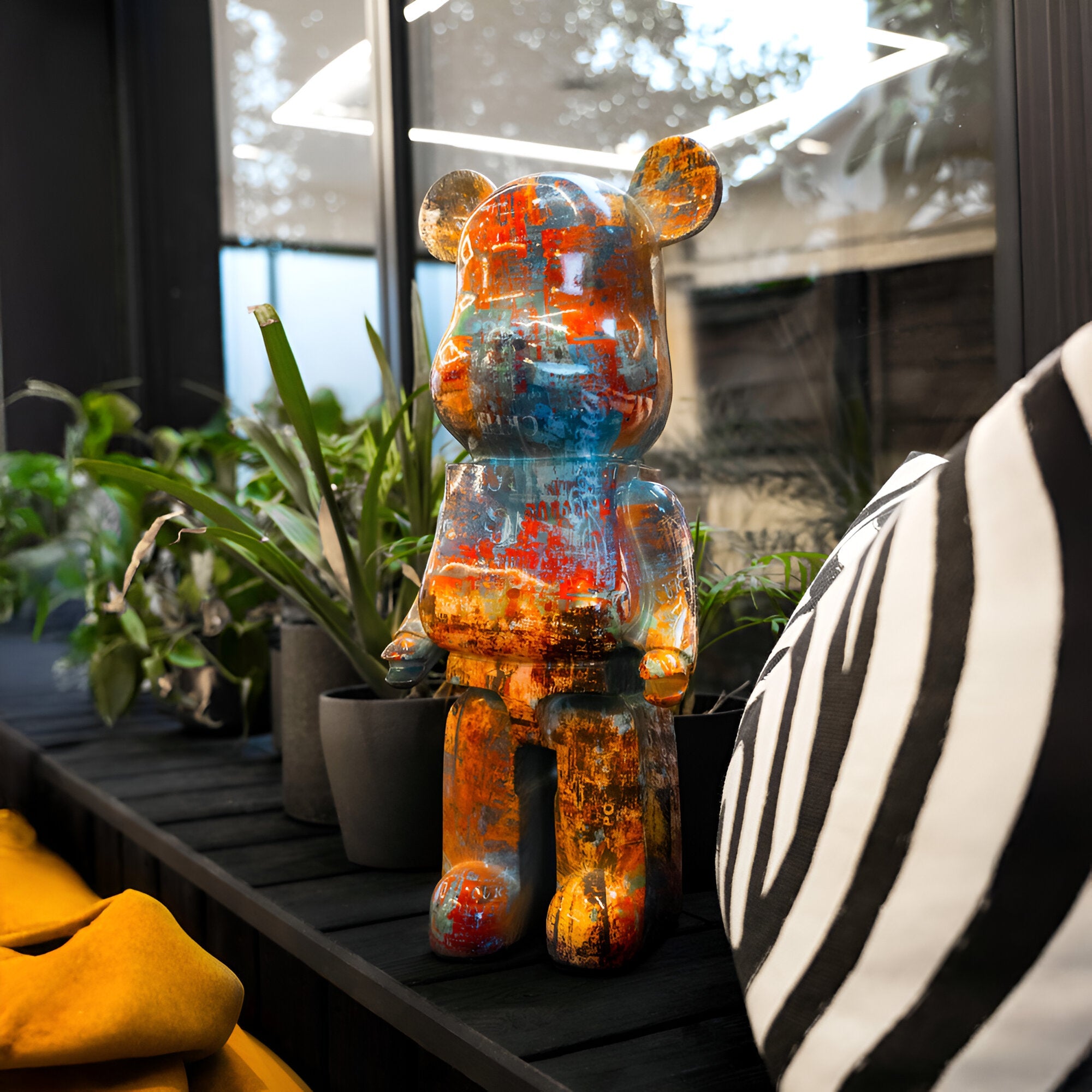 A vibrant and graffiti-inspired Obsidian Bear Sculpture by Giant Sculptures graces a table in a contemporary room. This 50cm artistic home decor piece is elegantly surrounded by potted plants, complemented by geometric lighting above and a zebra-striped cushion nearby, seamlessly integrating into the modern design.