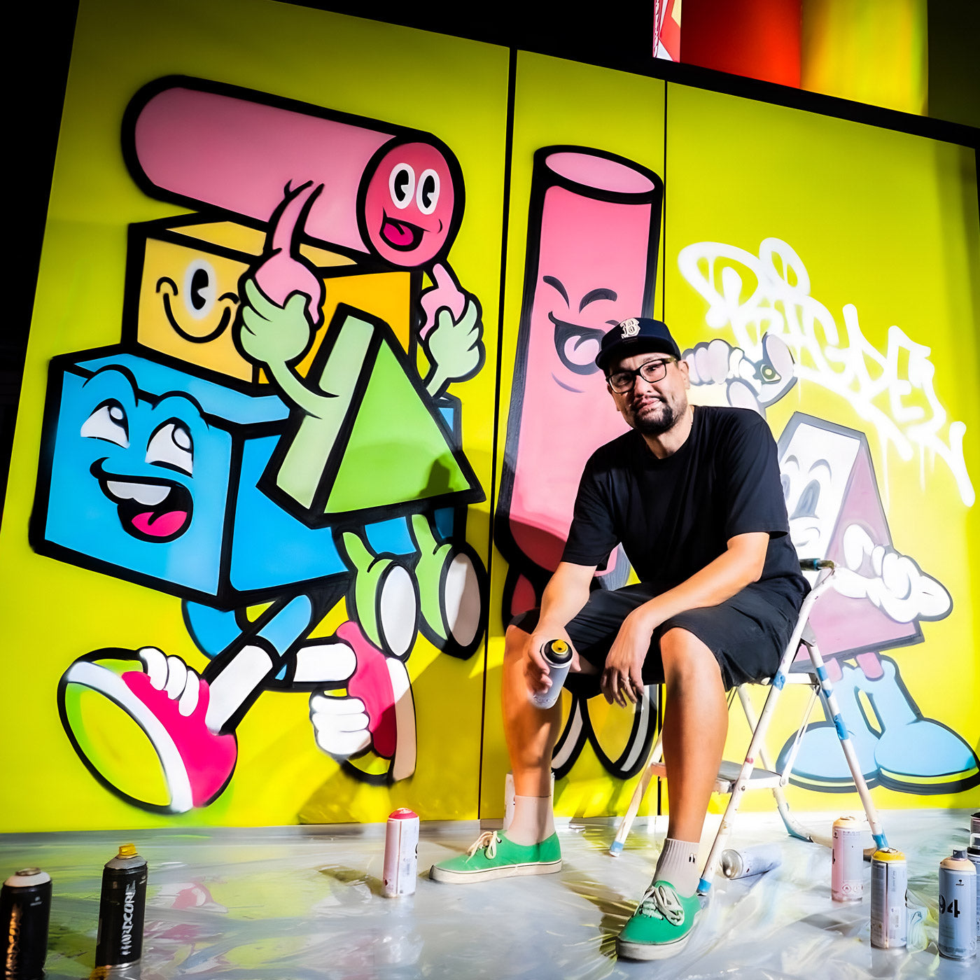 Artist sitting on a chair holding a spray can in front of a vibrant graffiti mural featuring colorful geometric characters with smiling faces. The wall is bright yellow, and several spray paint cans are scattered on the floor.