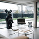 A spacious living room with expansive windows reveals a stunning outdoor view. Within the space, an elegant wildlife-inspired art piece takes center stage: the Black Stone Bear Sculpture - 165cm by Giant Sculptures. Completing the room's modern aesthetic are a sleek black chair, an understated side table, and a pristine white coffee table adorned with books. The wooden deck outside is framed by abundant greenery.