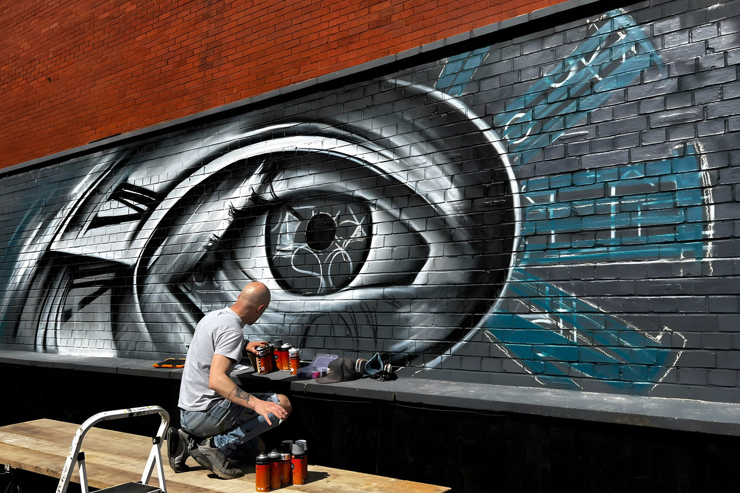 A person sits on a ladder in front of a large, detailed mural on a brick wall, featuring a stylized eye with blue and black accents. Several spray paint cans are placed on a wooden plank nearby.