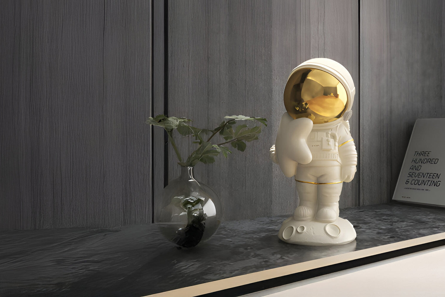 A white astronaut figurine with a shiny gold helmet stands on a dark marble surface. Next to it is a clear vase with a leafy plant. In the background, there is a framed picture with text partially visible on a wooden wall.