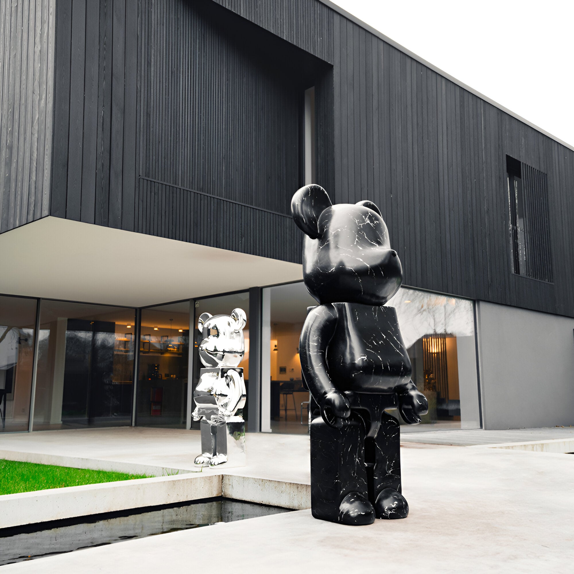 A modern, minimalist building with gray and wooden features showcases two wildlife-inspired bear sculptures from Giant Sculptures, including the Black Stone Bear Sculpture - 165cm and a silver counterpart. These statues stand by a reflecting pool under cloudy skies.