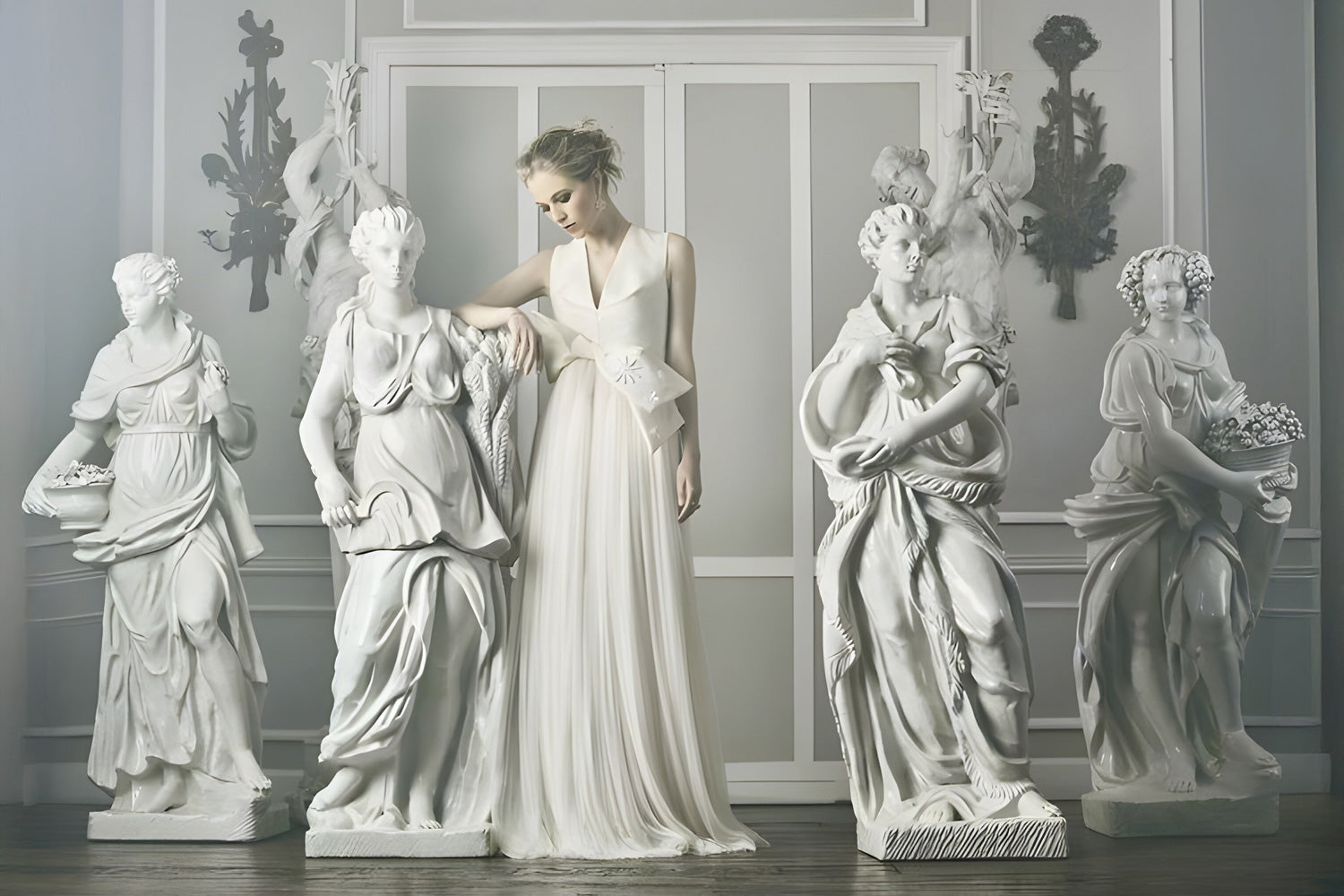 A woman in an elegant, flowing white gown stands among four white statues in a classical setting. She gently rests her hand on the shoulder of one statue. The scene is serene and ethereal, with soft lighting enhancing the graceful atmosphere.