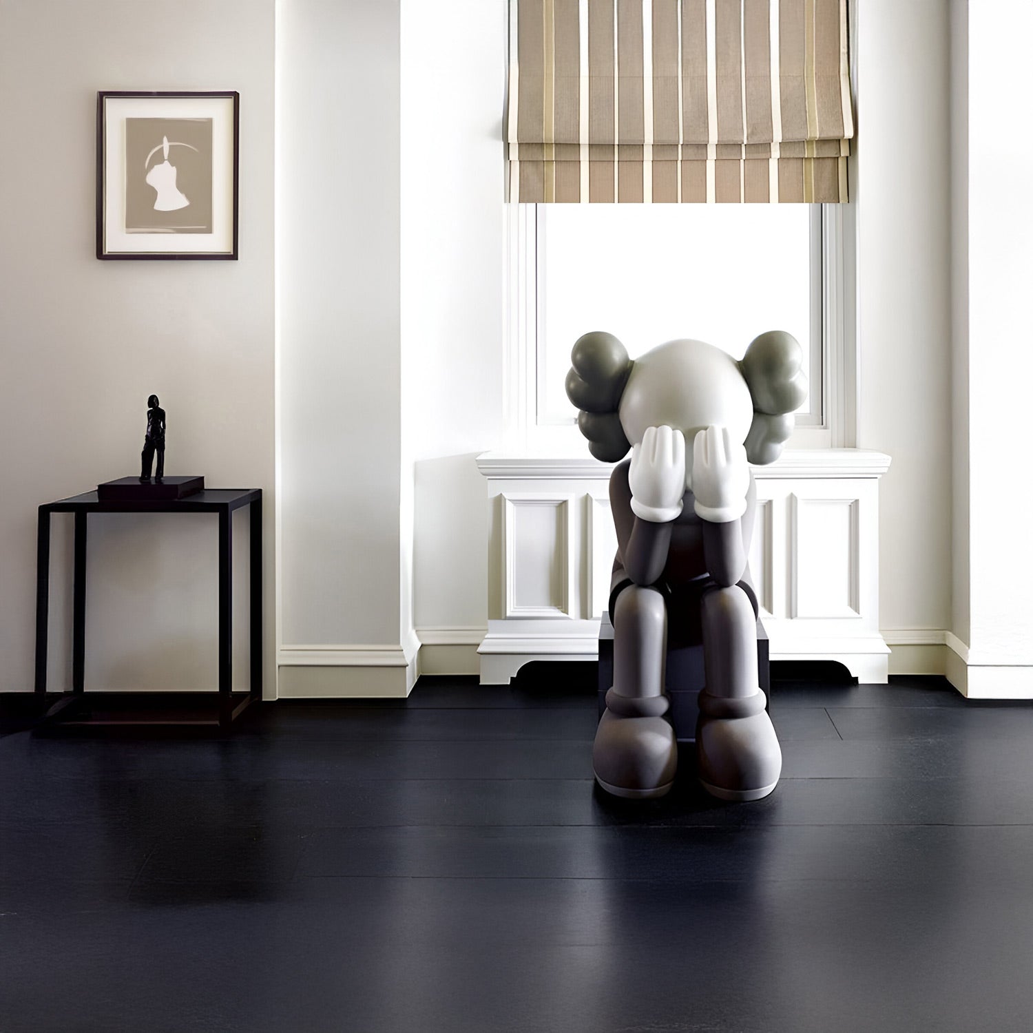 A modern room with a large gray figure sitting on the floor with its face in its hands. A small black table holds a statue and a framed picture hangs above it. Light filters through a striped window shade.