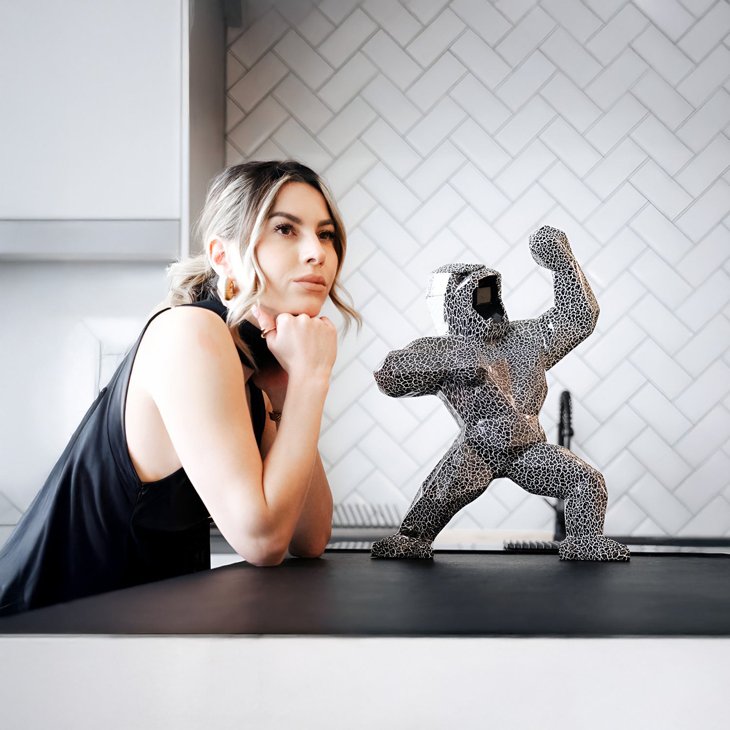 A woman with light hair leans her chin on her hands, gazing at a black and white sculpture of a person flexing muscles on a kitchen counter. The background features a herringbone-patterned tile wall.