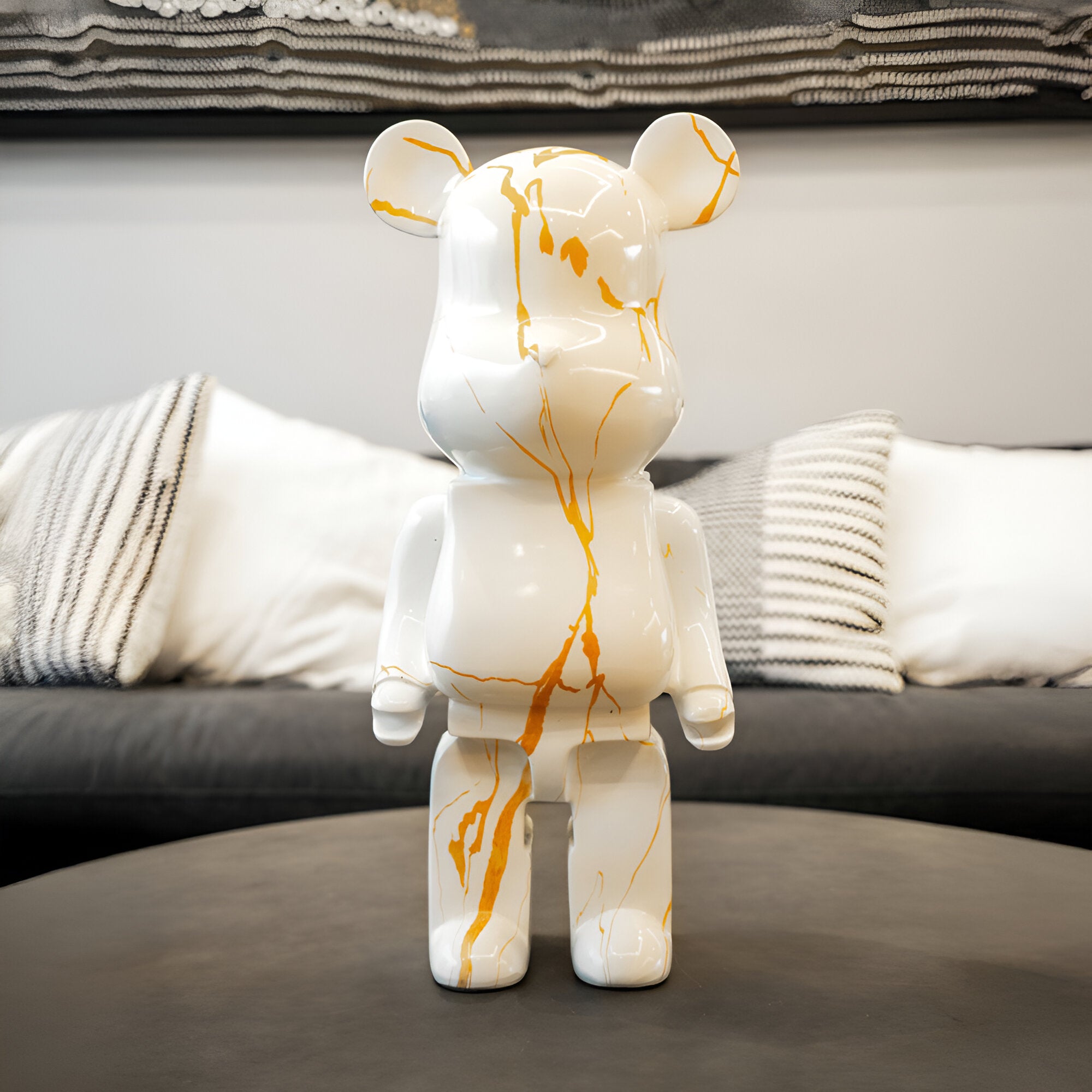 The "Golden Luster Bear Sculpture - 50cm" by Giant Sculptures, with its luxurious white and gold marbling design, stands elegantly on the table. In the background, a gray sofa is adorned with both striped and solid cushions, complemented by a piece of textured contemporary art on the wall.