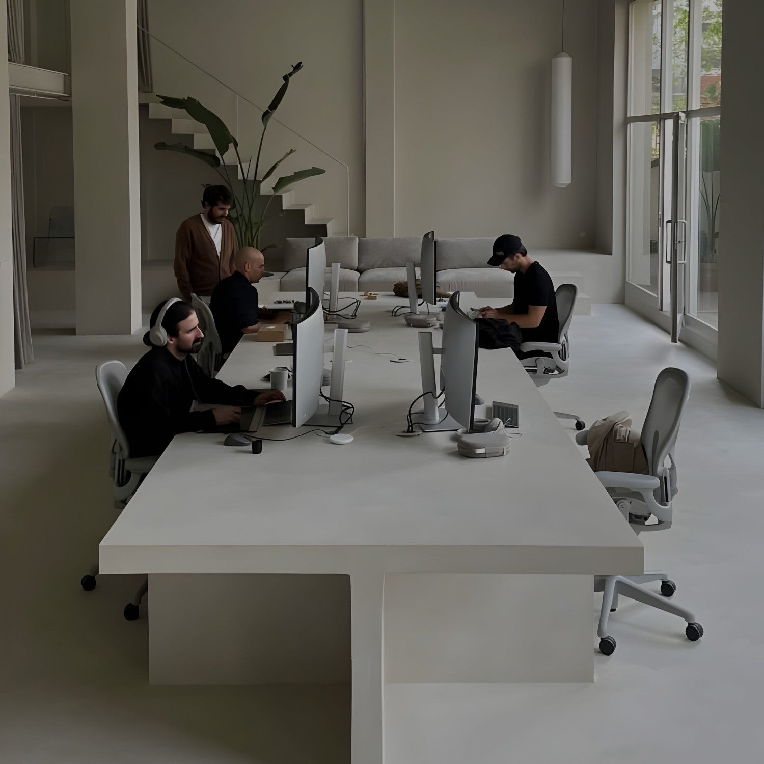 A modern office space with four people working at a sleek white table. They are using desktop computers and wearing casual attire. Large windows and minimalist decor, including plants and a staircase, create an airy atmosphere.