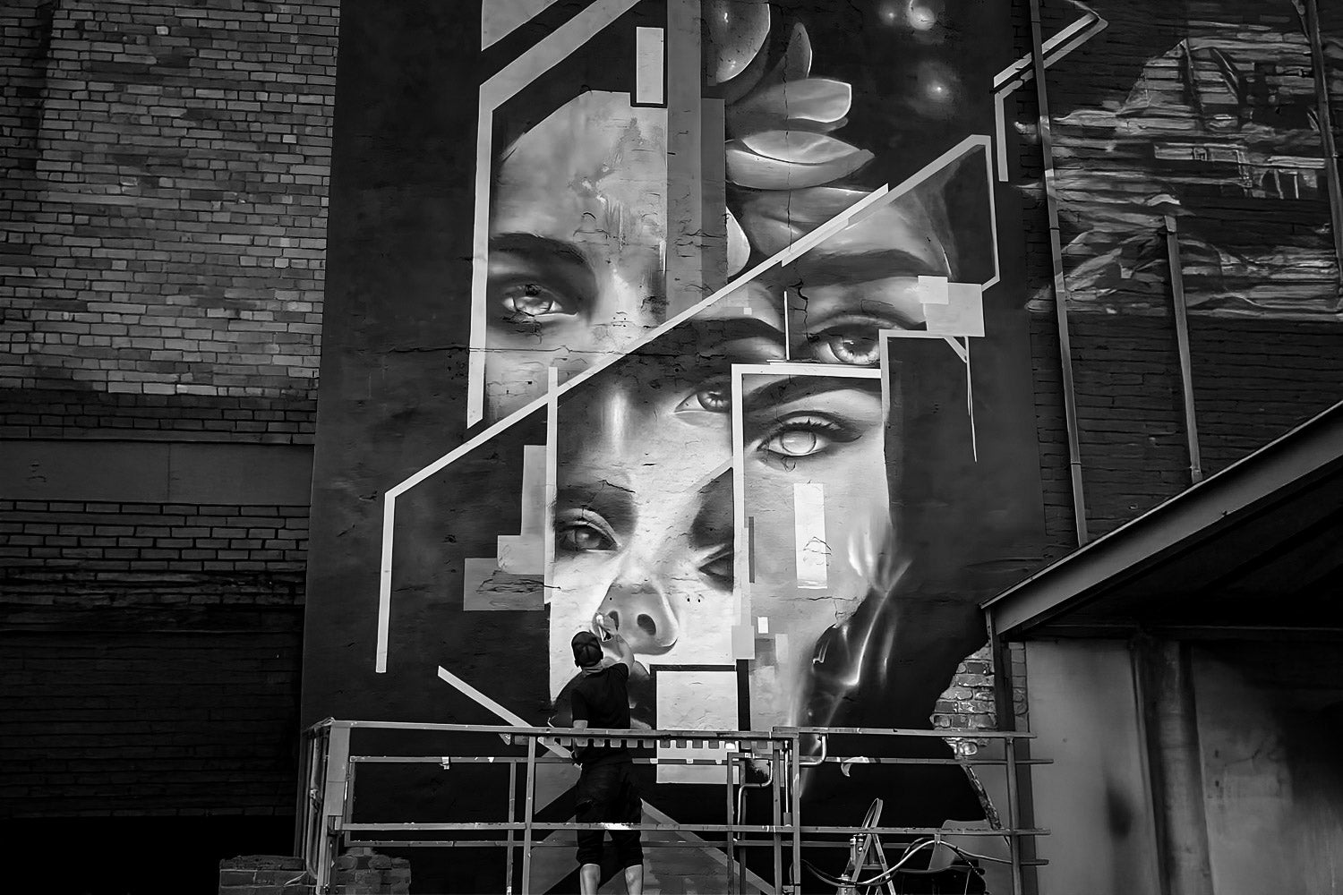 A person stands on scaffolding in front of a large black and white mural on a brick wall, featuring abstract, fragmented human faces with geometric elements.