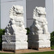 The Chinese Guardian Lion II Marble Outdoor Sculptures by Giant Sculptures consist of two large white stone statues on ornate square pedestals. Positioned outside in front of lush green shrubbery and a white building, these hand-carved lions feature intricate details and seem to protect the entrance.