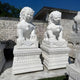 The Chinese Guardian Lion III Marble Outdoor Sculptures by Giant Sculptures, two large and intricately carved white stone statues perched on ornate pedestals, stand majestically against a backdrop of a white brick wall beneath a clear blue sky.