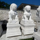 Two large, hand-carved Chinese Guardian Lion III Marble Outdoor Sculptures by Giant Sculptures sit majestically on intricately carved pedestals. The clear blue sky highlights the detailed designs against a white brick wall.