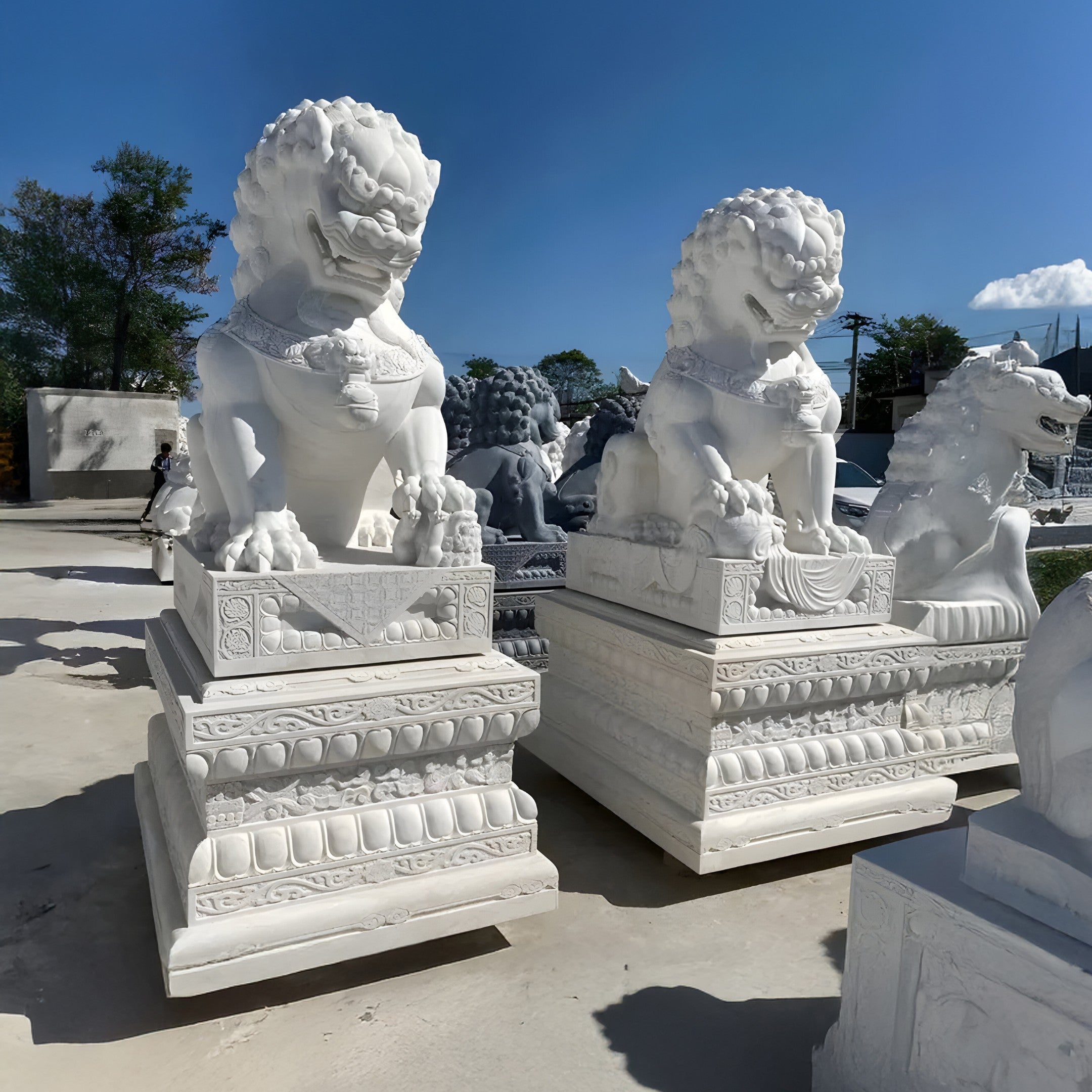 Under a clear blue sky, the **Chinese Guardian Lion III Marble Outdoor Sculpture** by **Giant Sculptures** majestically sits atop ornate pedestals. Sunlight accentuates the intricate carvings on both the pedestals and the statues, while additional stone sculptures can be seen in the background.