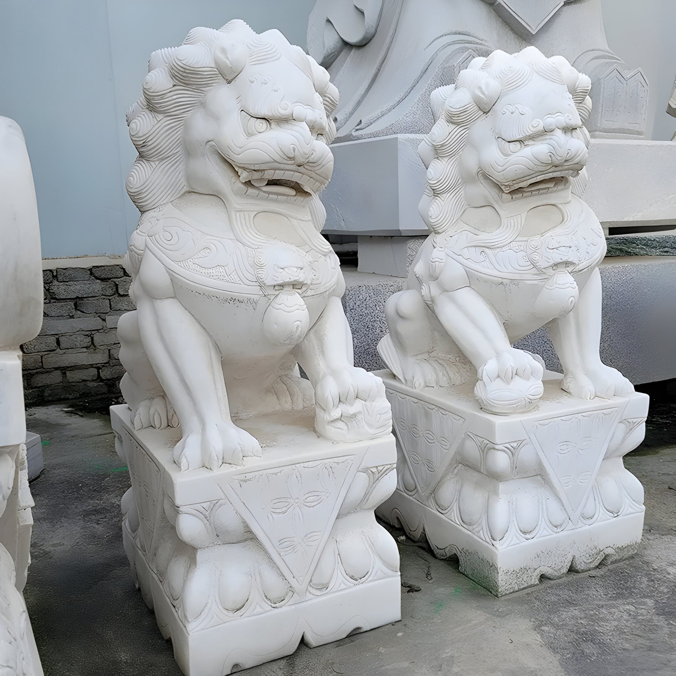 Placed side by side, the "Chinese Guardian Lion IV Marble Outdoor Sculptures" from Giant Sculptures stand majestically on ornate pedestals, exemplifying exquisite marble craftsmanship. Each lion features a meticulously detailed mane and a fierce expression. In the background, other sculptures can be seen partially obscured.