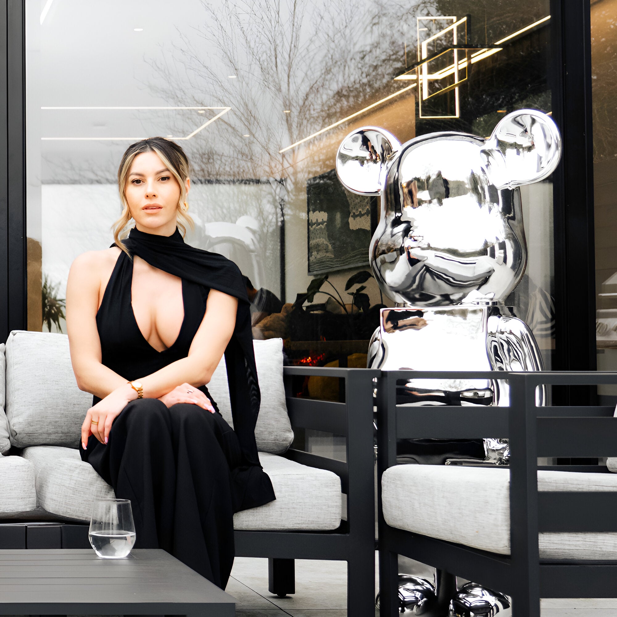 A woman in a black dress sits on a modern outdoor sofa next to Giant Sculptures' Mystic Chrome Bear Sculpture - 135cm, its chrome electroplated finish gleaming. The setting features a stylish patio with reflective glass doors and minimalistic decor, representing the epitome of modern design.