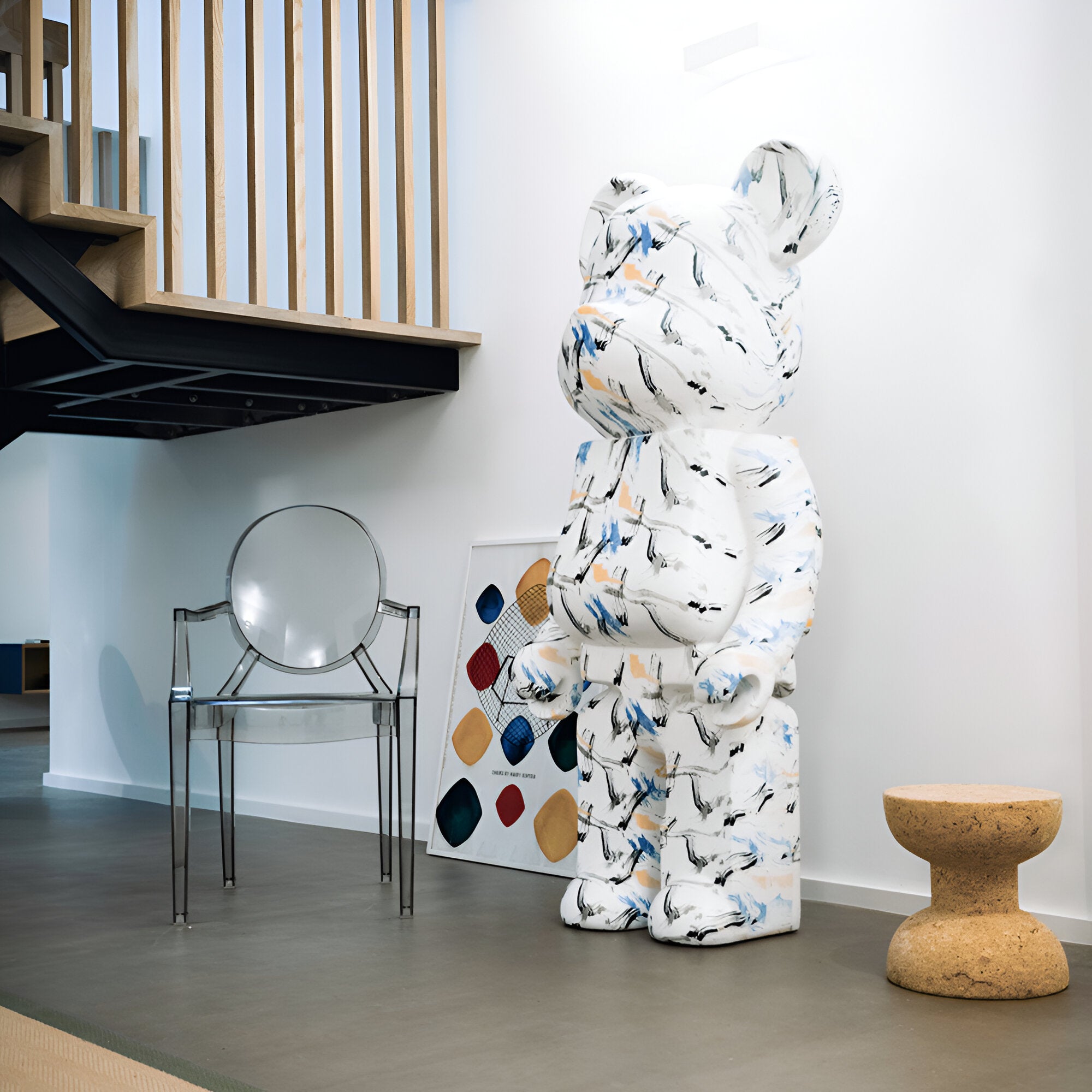 A contemporary interior space features the eye-catching Indigo Infinity Bear Sculpture - 165cm by Giant Sculptures, designed with a marble pattern. The sculpture is paired with a clear chair and a cork stool for seating. In the background, an abstract artwork presents vibrant geometric shapes, and on the left side of the room, you can partially see a wooden staircase.