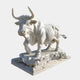 The Charging Bull White Marble Outdoor Sculpture - 150cm by Giant Sculptures features a detailed bull with strong features and raised front leg on a decorated base, highlighting its texture and musculature.