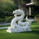 The 120cm Imperial Dragon White Marble Outdoor Sculpture by Giant Sculptures commands attention with its intricate scales and fierce expression. It stands proudly on the manicured lawn, set against a serene garden, lush bushes, and a traditional building backdrop.