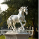 The Galloping Horse Marble Outdoor Sculpture - 160cm by Giant Sculptures depicts a lifelike horse in mid-gallop with a flowing mane and tail, proudly mounted on a decorated pedestal amid an outdoor park setting, framed by trees.