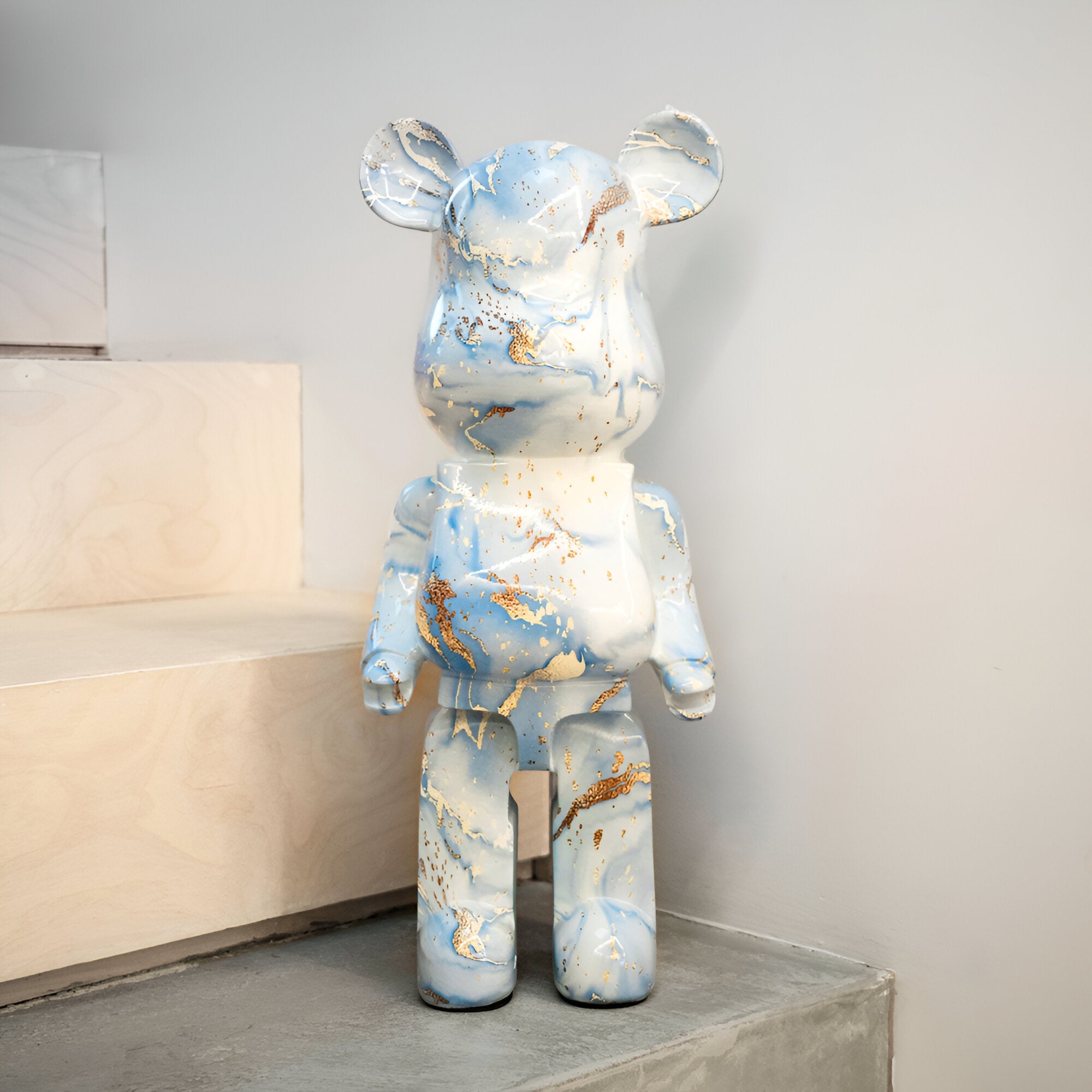 The Ocean Spray Bear Sculpture - 50cm from Giant Sculptures features a marbled blue and white finish accented with gold flecks, evoking the fluidity of waves. Positioned on a step bordered by light-colored stairs and walls, it embodies a minimalist and modern style that complements any contemporary bear sculpture collection.