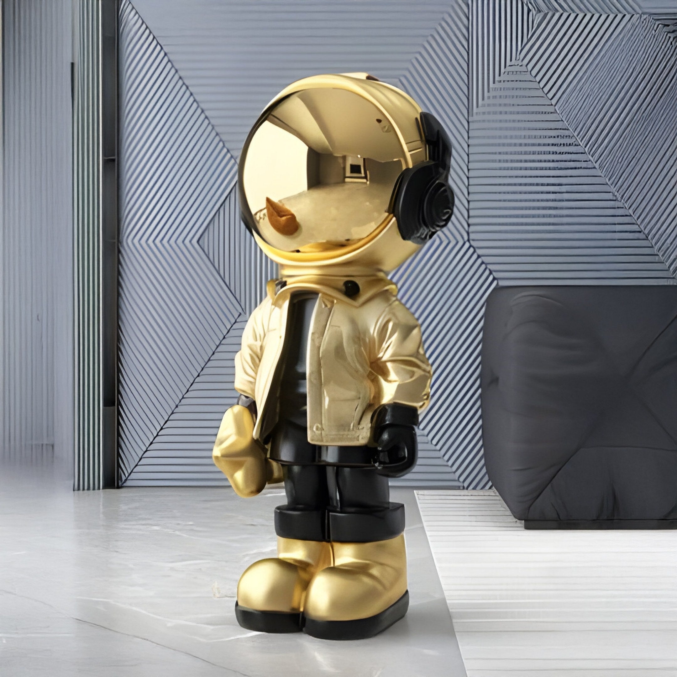 The Celestial Gold Astronaut Beats Sculpture by Giant Sculptures stands on a patterned white and gray floor, featuring a shiny gold helmet, jacket, and boots. With a backpack against a geometric wall backdrop, this 100cm resin figure embodies a space-inspired theme.