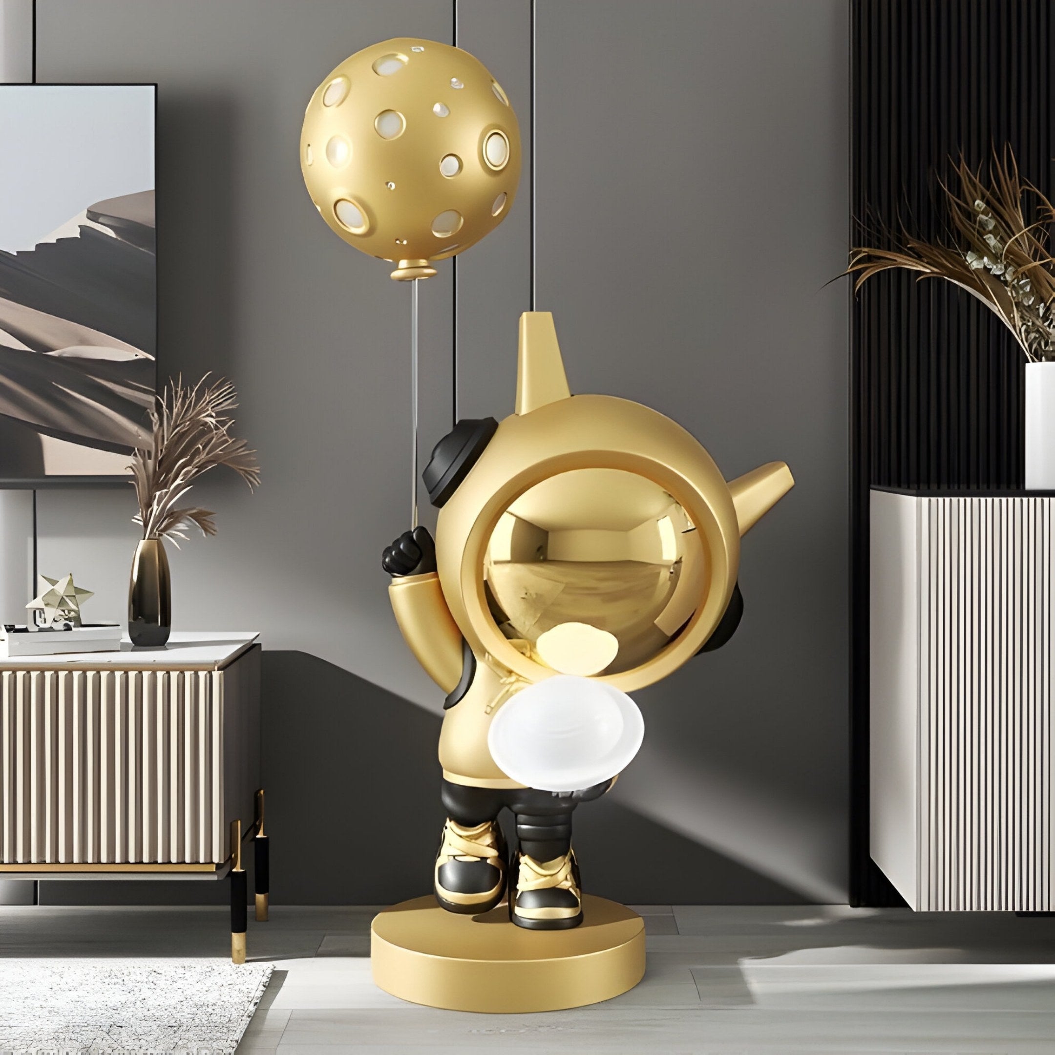 The Celestial Gold Astronaut DJ Moon Balloon Sculpture (100cm) by Giant Sculptures adds a space-inspired touch to a modern room. The shiny golden figure with a reflective helmet and black gloves stands elegantly among decorative vases and geometric walls.