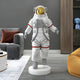 A modern living room boasts the White Space Astronaut Voyager Sculpture - 100cm by Giant Sculptures, featuring a life-sized astronaut with a gold visor. The space includes a gray couch with orange pillows, a beige bean bag, and shelves filled with decorative items, all evoking space exploration spirit.