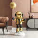 In a modern room with a beige armchair, black and white cabinet, and warm-toned abstract art, the Cosmic Gold Astronaut Moon Balloon Sculpture by Giant Sculptures stands on a pedestal.