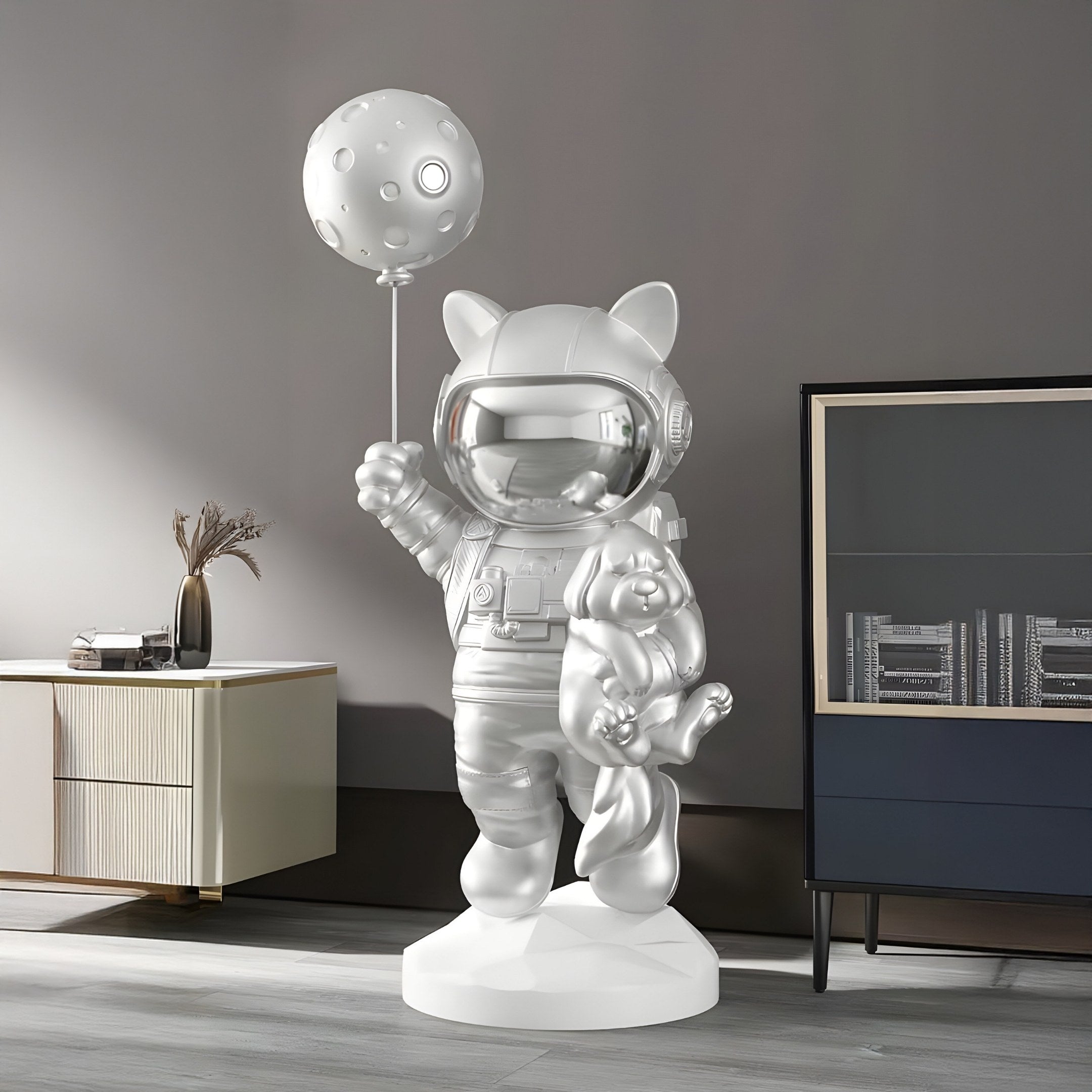 The Cosmic Chrome Astronaut Kitty Pup Moon Balloon Sculpture (110cm) by Giant Sculptures adds whimsy to a modern room with its futuristic design, featuring an astronaut kitty holding a moon balloon and teddy bear against a minimalistic gray wall.