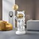 In a modern room, the Nova White Astronaut Kitty Pup Moon Balloon Sculpture by Giant Sculptures stands whimsically on a pedestal. It features cosmic charm against sleek, neutral-toned walls with a yellow beanbag and lamp nearby.