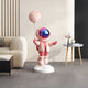 The Giant Sculptures Stellar Pink Astronaut Moon Balloon Sculpture, 110cm, features a shiny visor and pink balloon, standing on a white base beside a beige sofa and simple side table with clock and vase. It complements walls with vertical lines for whimsical décor.